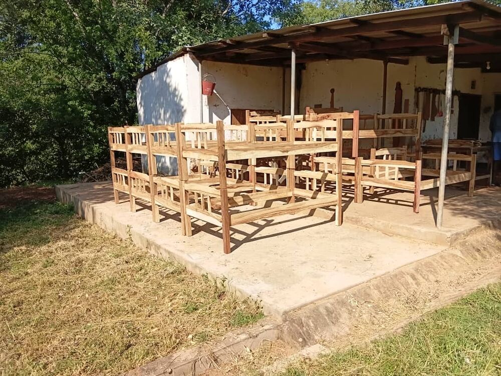 Several unfinished wooden bunk beds and single beds are arranged under a simple outdoor shelter