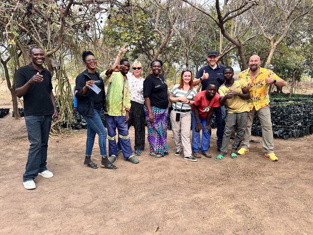 A group of eleven people, including adults and youths, smilingly poses in front of trees and plant bags on dirt ground