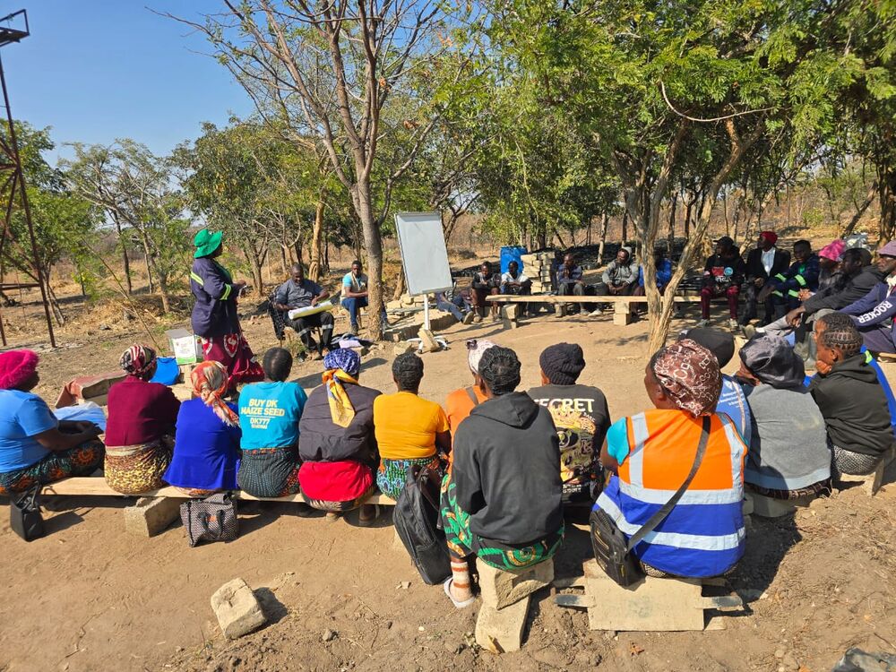 A group of women and men sit outdoors under trees, listening to a woman speaking while a man takes notes