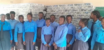 A group of students in blue uniforms stands in front of stacked mattresses in a classroom