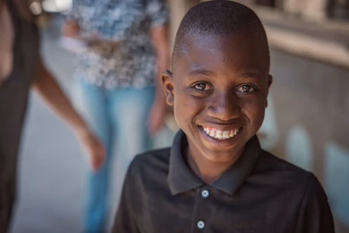 A smiling boy with dark skin and short hair looks directly into the camera, blurred people in the background