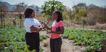 Two women converse in a vegetable garden with rows of cabbage, one gesturing while holding keys and plant material, the other listening