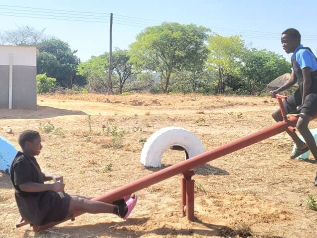 Two children play on a seesaw made of metal and tires in a dry playground with a building and trees in the background