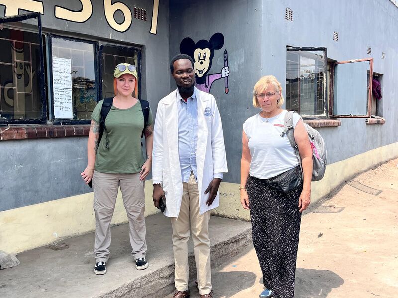 Three people, two women and a man in a lab coat, stand in front of a light blue building with large numbers "4567" and a Mickey Mouse drawing