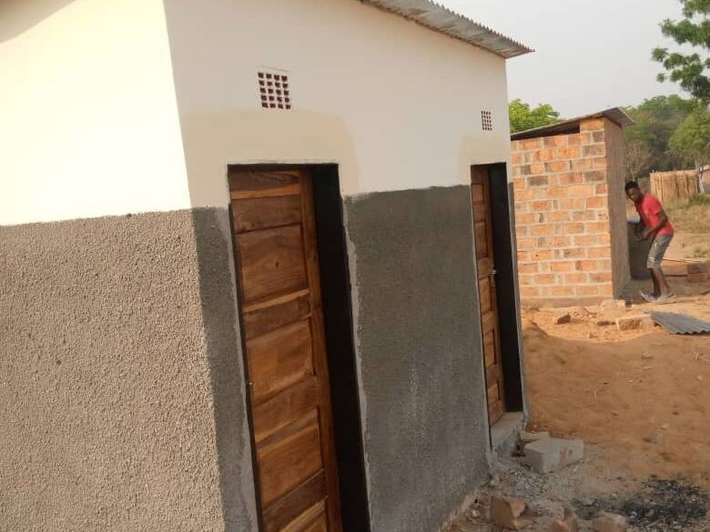 Newly constructed toilet building with two wooden doors and a corrugated metal roof, a man is working on another structure in the background