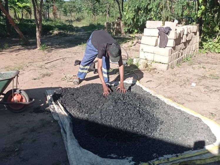 A man processes a large pile of dark, granular material, likely charcoal, spread on a tarp outdoors