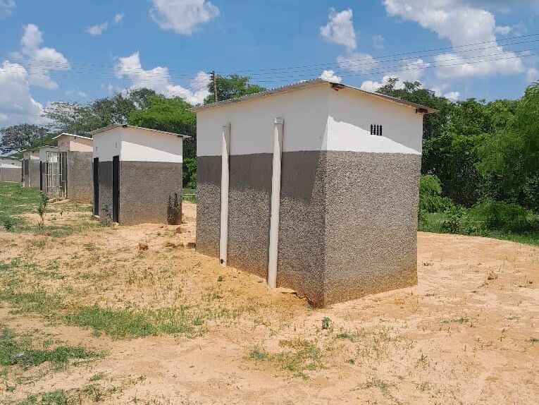 Row of simple, rectangular buildings with two-tone facades and pipes on dry, sandy ground under a blue sky