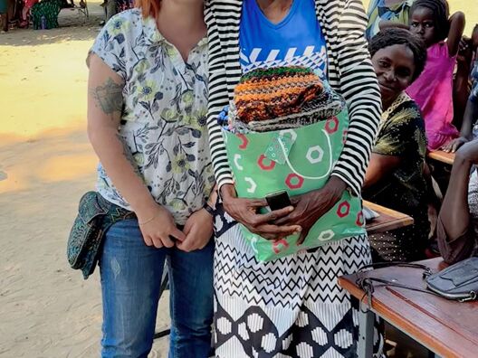 Two women smiling at the camera, one holding a colorful bag. Other people sitting in the background under a tree.