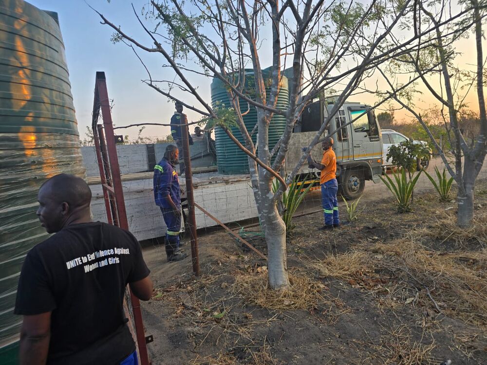Several men work outdoors near a truck and large water tanks, one wearing a t-shirt with text