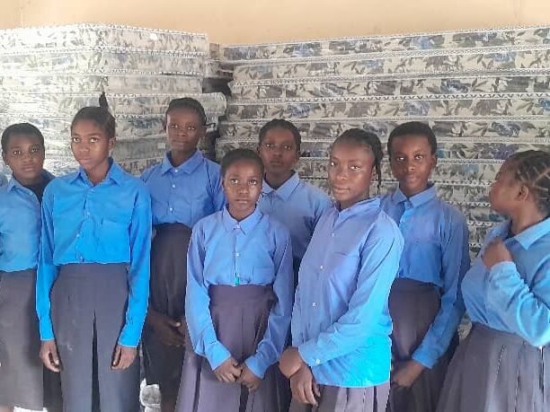 A group of students in blue uniforms stands in front of stacked mattresses in a classroom