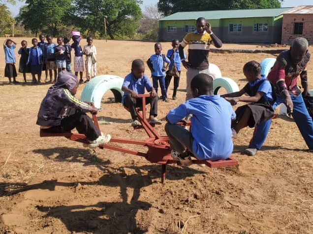 Children play on a dusty schoolyard with a seesaw-like merry-go-round and painted tires, other children stand in the background
