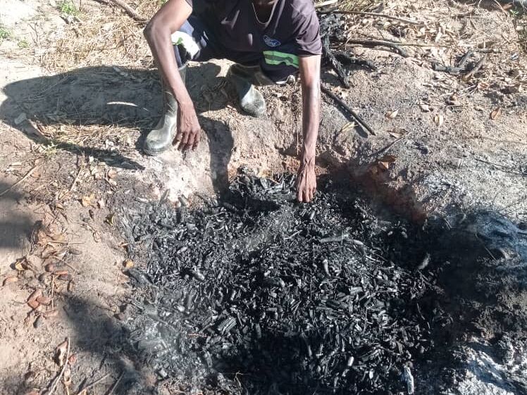 A man crouches over a pile of charcoal and ash on the ground, working with it using his hands