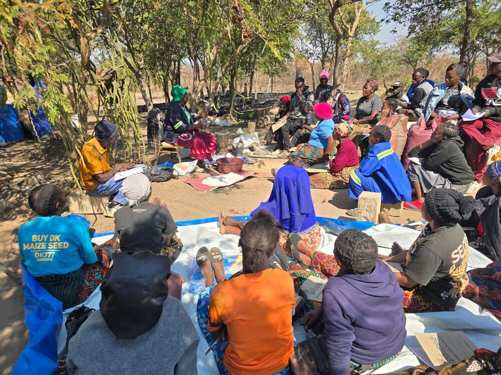 A group of people, mostly women, sitting outdoors in a circle under trees, attending a meeting or training session