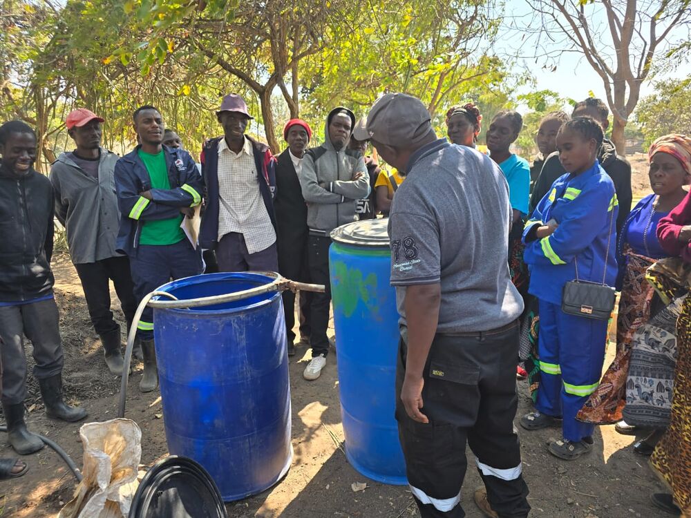 A group of people stands outdoors around two blue barrels, with one person facing away from them