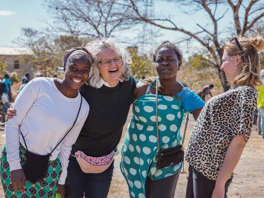 Four women of diverse ages and backgrounds stand closely together, smiling and laughing in a sunny outdoor setting