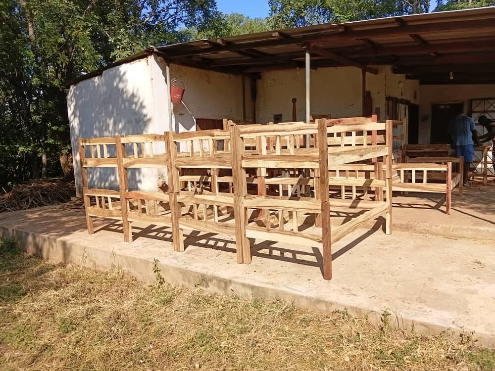 Several new, unvarnished wooden bunk beds are placed in front of a simple building, with people working in the background