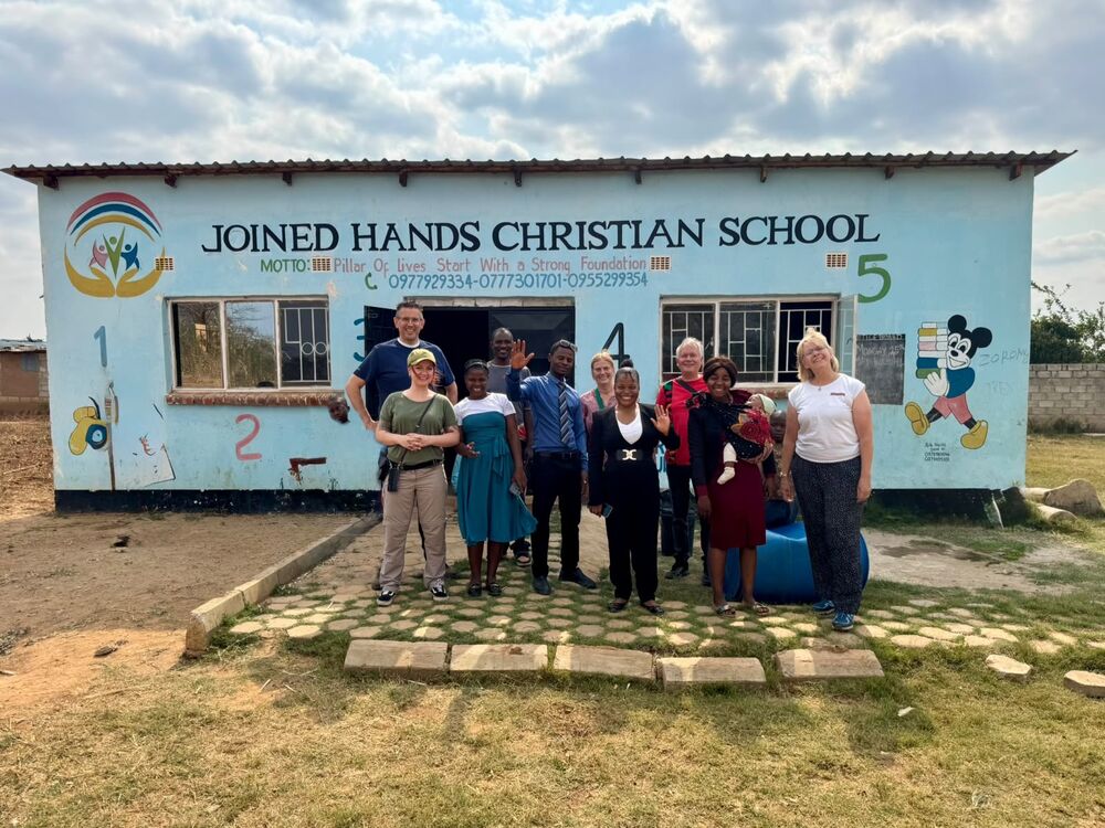 A group of adults and a baby stand smiling and waving in front of the blue "Joined Hands Christian School" building