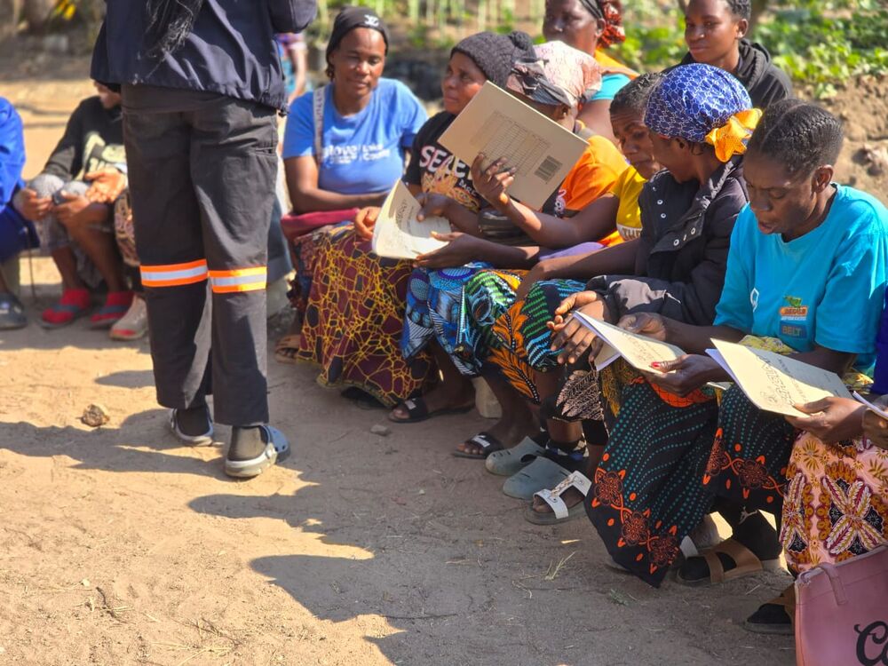 A group of women sits outdoors, holding papers or notebooks, while a person in reflective trousers stands in front