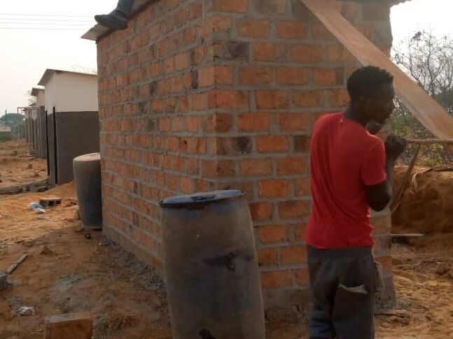 Two men constructing a small brick hut; one sits on the roof with corrugated metal, the other stands on sandy ground