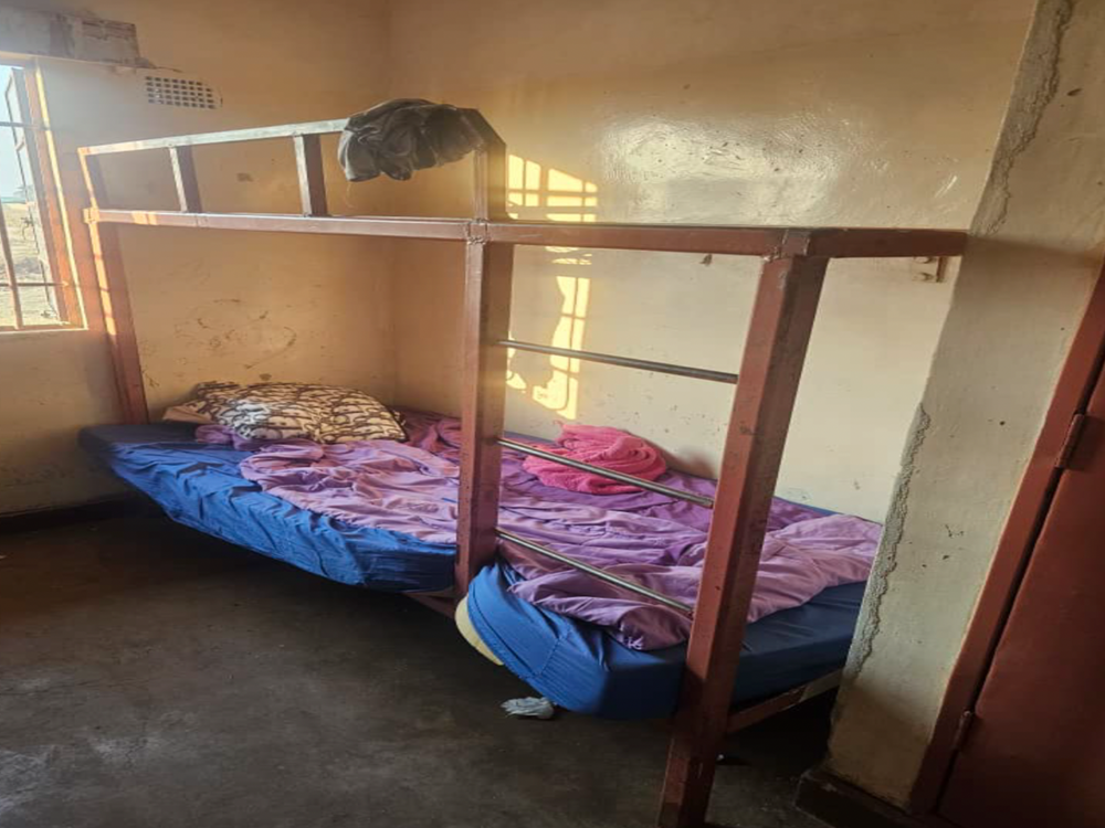 A simple room featuring a metal bunk bed with a blue mattress and purple bedding on the lower bunk, sunlight streams through a window