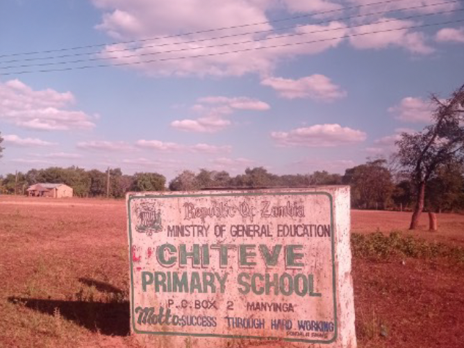 A weathered sign for Chiteve Primary School in Zambia stands on dry, reddish ground under a blue sky