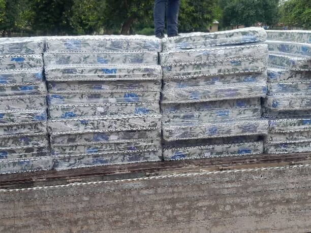 Numerous plastic-wrapped mattresses with a floral pattern are stacked high, a person's legs are visible on top