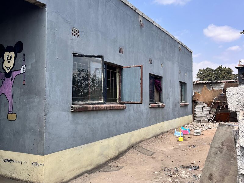 Grey building with yellow base and Mickey Mouse mural, in front a dusty yard with debris and children's toys under a blue sky
