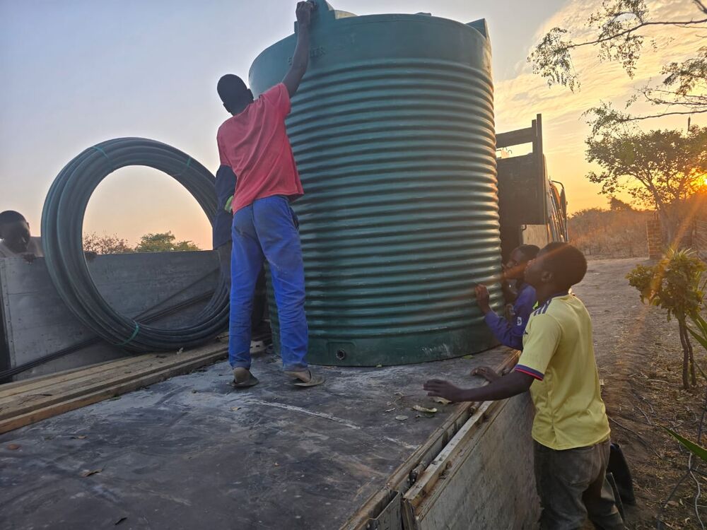 Several people load a large green water tank and black pipes onto a truck bed at sunset