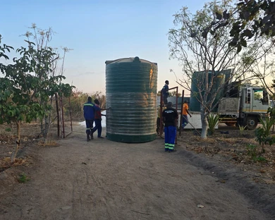 Several people are moving or installing large green water tanks outdoors, one tank is in the foreground, another on a truck