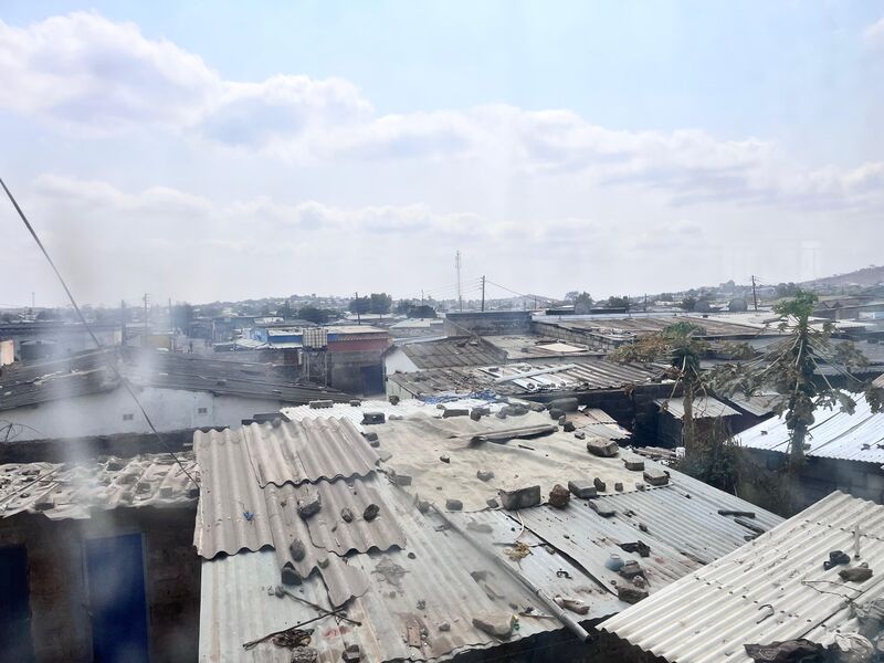 View over a densely populated area with many corrugated iron roofs, smoke rising, hills and clouds in the background