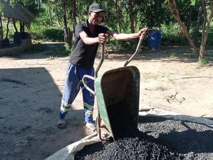 A man in a cap and work clothes empties a wheelbarrow full of black granular material onto a sack outdoors