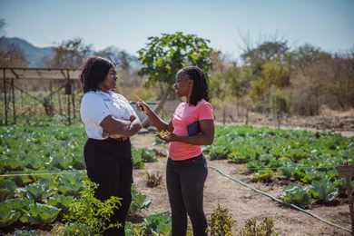 Two women converse in a vegetable garden with rows of cabbage, one gesturing while holding keys and plant material, the other listening