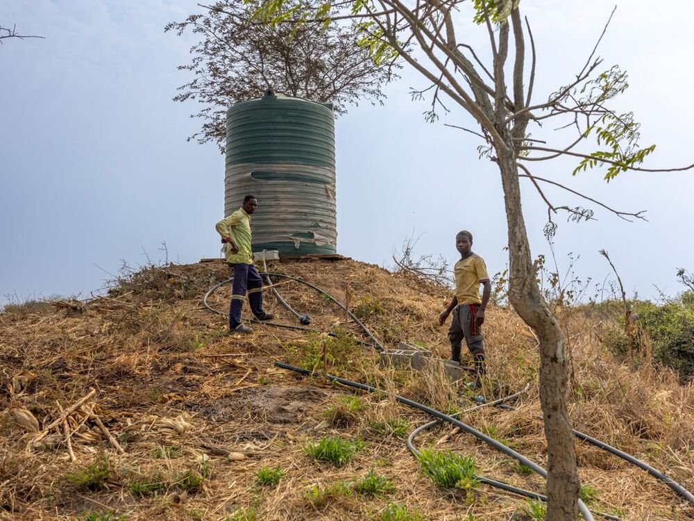Two men stand on a dry hill with a large green water tank and black irrigation pipes on the ground