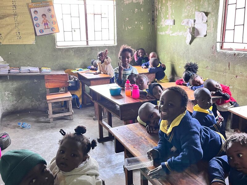 Many children in a simple classroom, some smiling at the camera, an older child holds a baby in the foreground