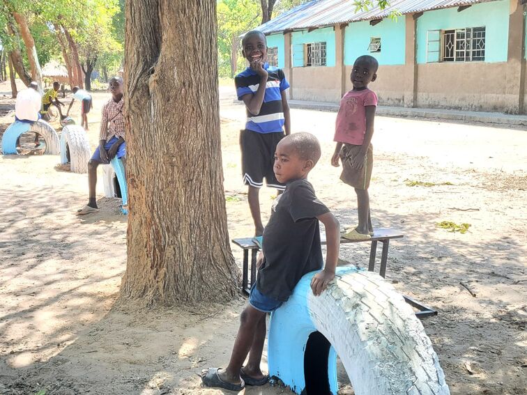 Children play on a dusty ground with painted tires and benches, a building and trees are visible in the background