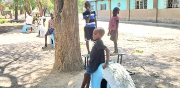 Children play on a dusty ground with painted tires and benches, a building and trees are visible in the background