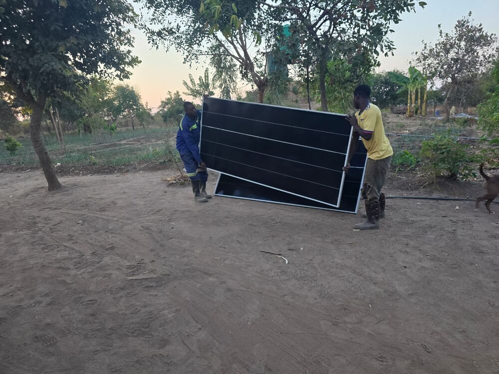 Two men are carrying large black solar panels across a sandy ground in a rural setting
